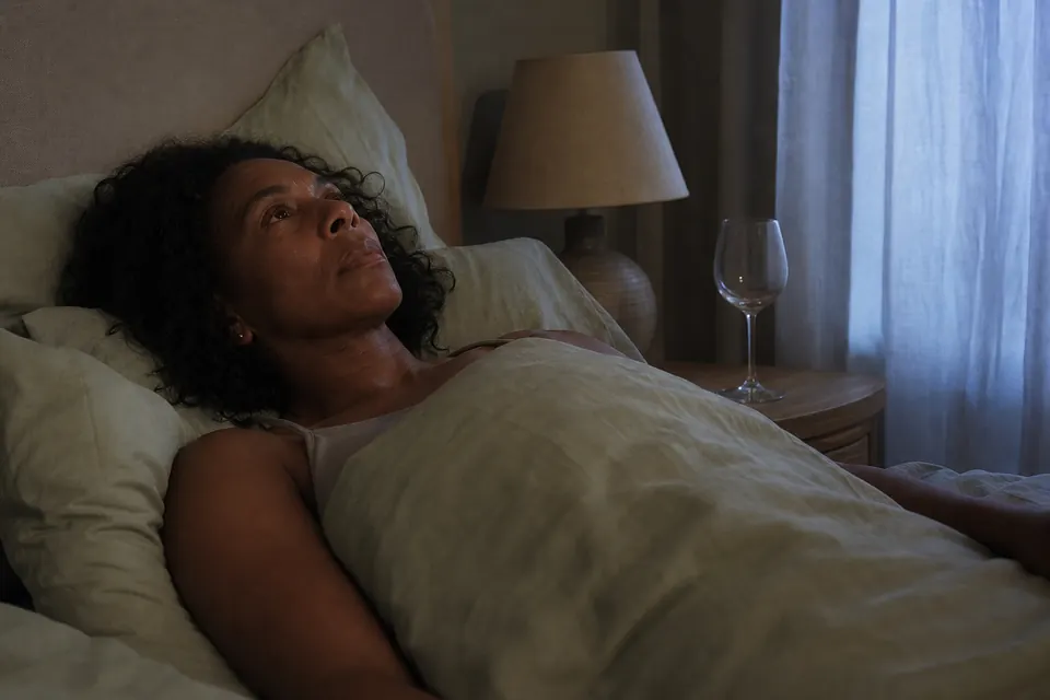 A Black British woman in her 40s lying awake in bed before dawn, an empty wine glass on her bedside table.