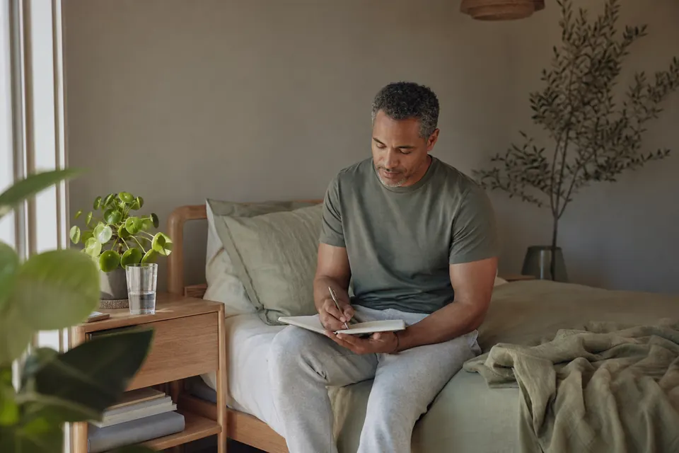 A mixed-race man in his 40s sitting on his bed in morning light writing a short note in a notebook.