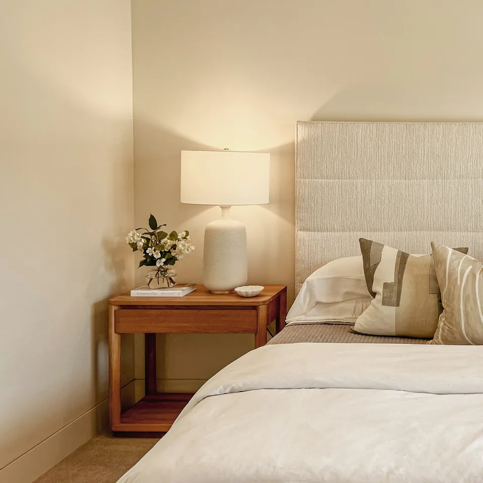 A softly lit bedroom with linen bedding, a wooden bedside table and flowers.