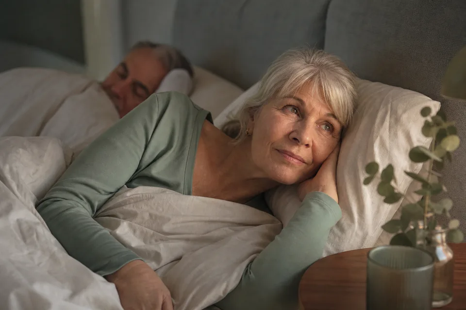 An older adult lies awake in a softly lit bedroom while a partner sleeps in the background.