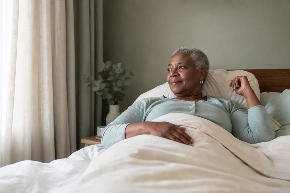 An older adult resting in bed as soft morning light enters the room.