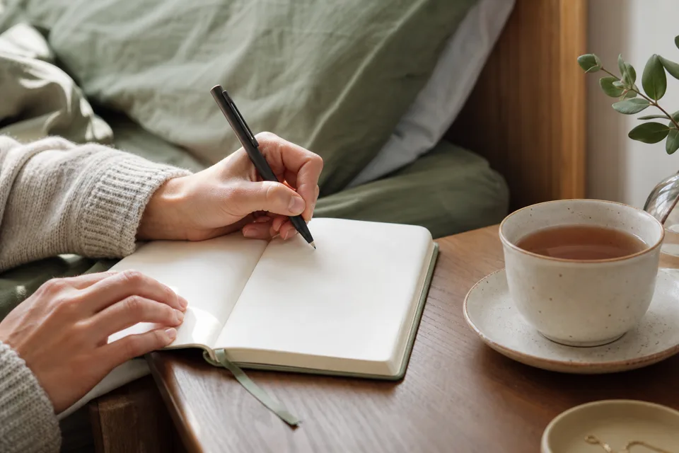 Hands making a short morning note in an open notebook beside a cup of tea.
